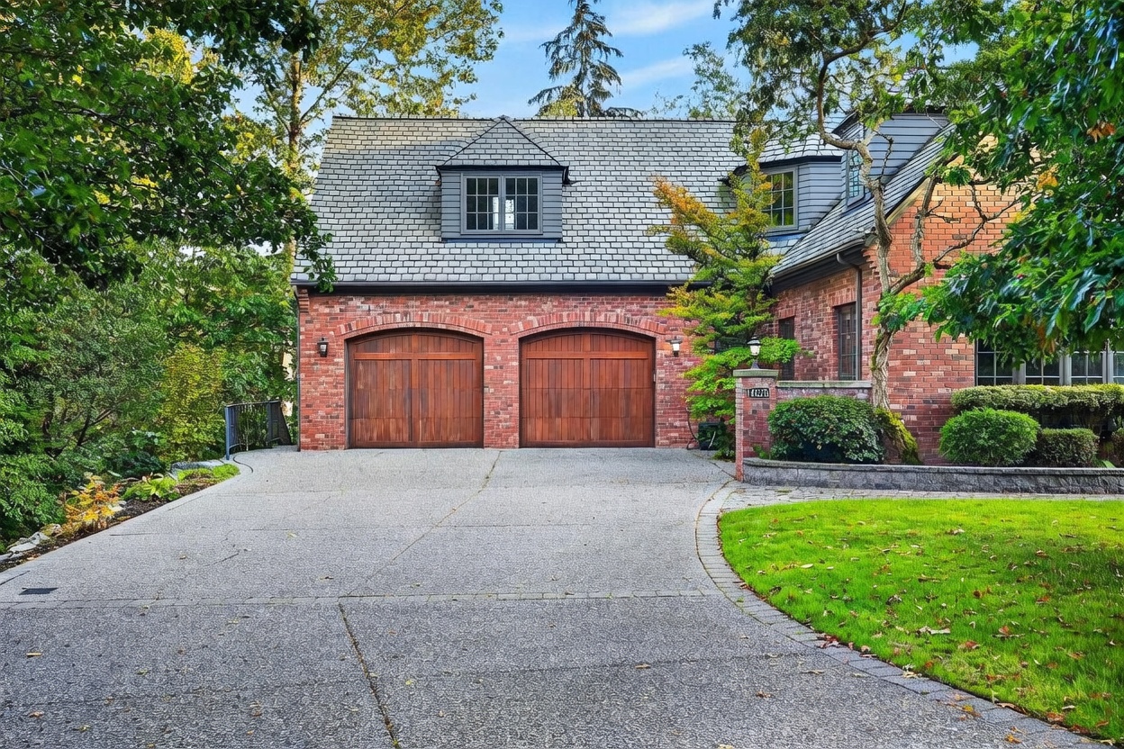 This image captures the front exterior of a traditional brick home, focusing on a prominent two-car garage with arched wooden doors. The structure features a steep, dark-shingled roof with a dormer window, complemented by lush green trees and a well-manicured lawn. A paved driveway leads up to the garage, creating a welcoming and established curb appeal.