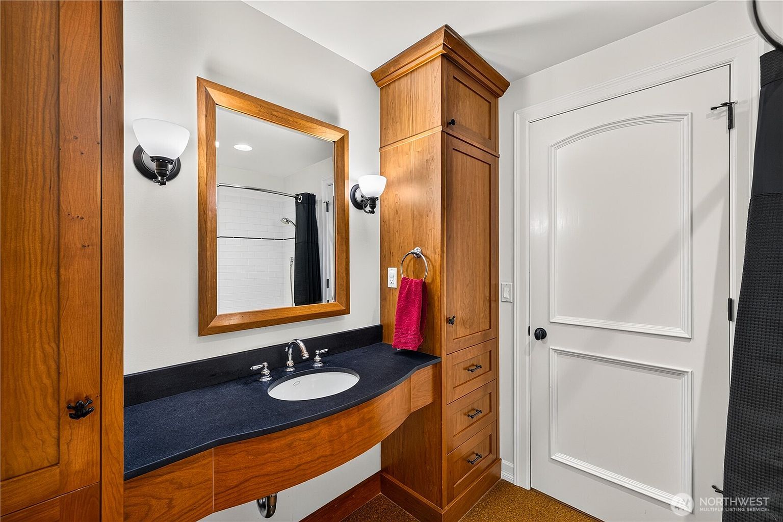 This bathroom features a warm, wood-toned vanity with a dark, contrasting countertop and an integrated sink. A framed mirror is centered between two wall-mounted light fixtures, while a tall, built-in storage cabinet provides additional organization. The space is finished with a white door and neutral wall tones, creating a clean and functional aesthetic.