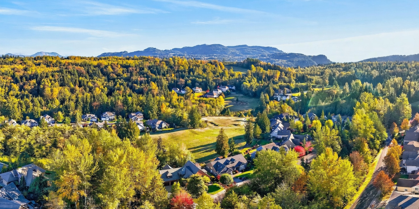 This high-angle aerial view captures a sprawling residential neighborhood nestled within a lush, forested landscape during the autumn season. The scene features multiple homes with varied rooflines surrounded by vibrant, color-changing trees and open green spaces. In the distance, rolling hills and mountains provide a scenic backdrop under a clear blue sky.
