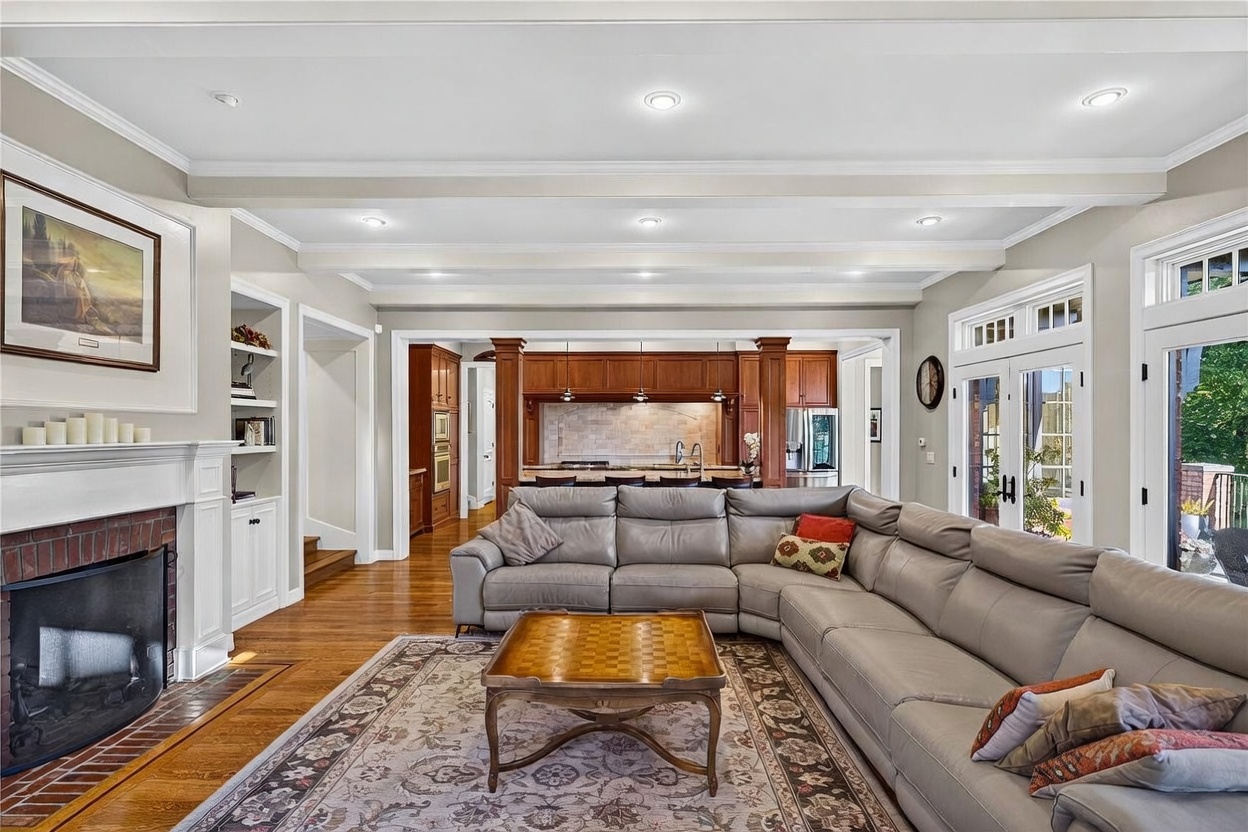 This spacious living room features a large, light-colored sectional sofa arranged around a wooden coffee table on a patterned area rug. To the left, a fireplace with a white mantel and brick surround serves as a focal point, flanked by built-in shelving. The room flows openly into a kitchen area with dark wood cabinetry and a breakfast bar, all illuminated by recessed lighting and natural light from the adjacent glass doors.