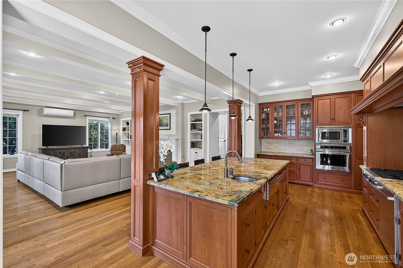 This open-concept kitchen features a large central island with a stone countertop and a built-in sink, illuminated by three pendant lights. The space is anchored by warm-toned wood cabinetry and stainless steel appliances, including a double wall oven. The kitchen flows seamlessly into a living area with a large sectional sofa, creating an inviting and spacious atmosphere.