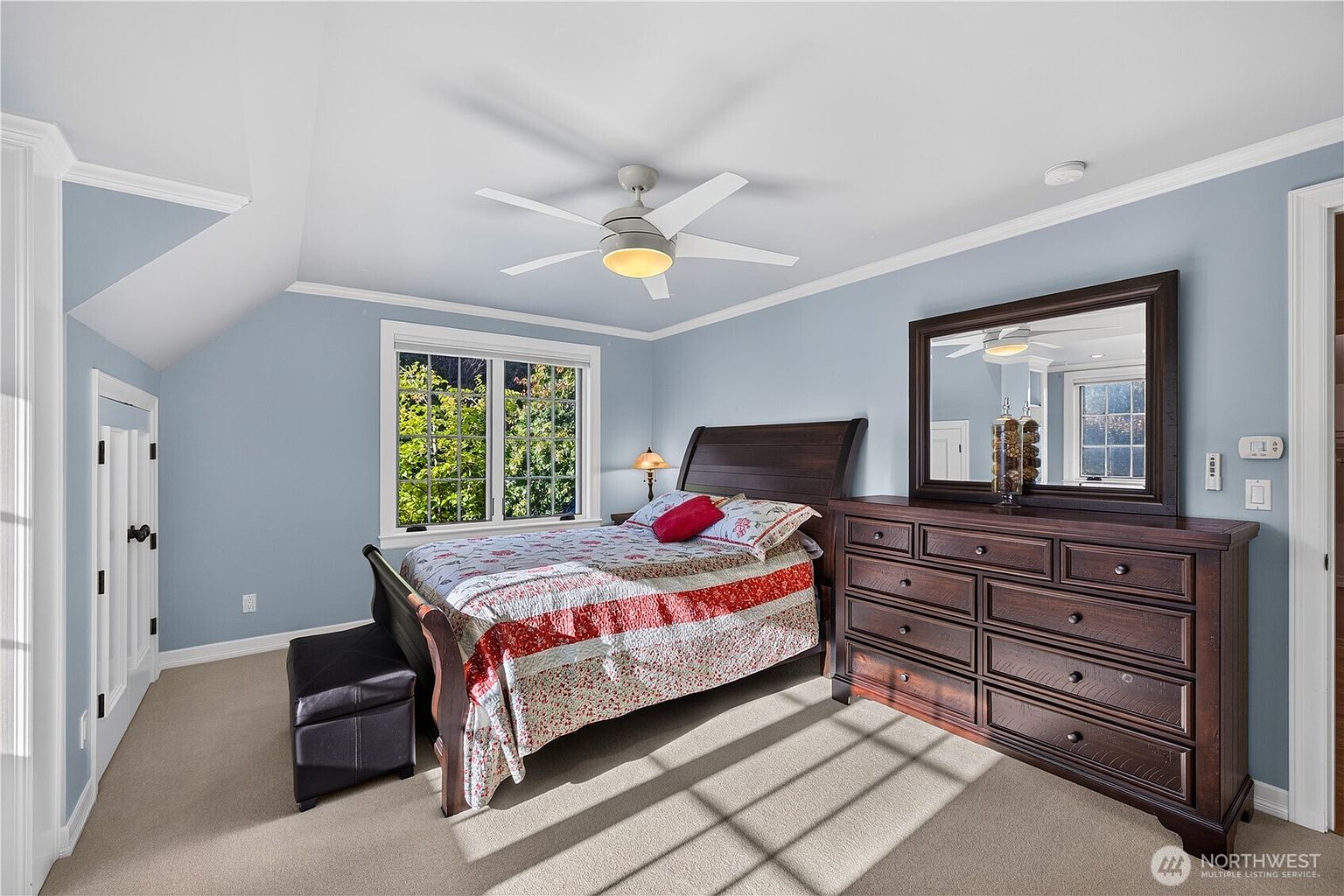This bedroom features light blue walls, neutral carpeting, and a dark wood bed frame paired with a matching dresser and mirror. A ceiling fan with integrated lighting provides overhead illumination, while a window allows natural light to brighten the space. The room is furnished with a small bench at the foot of the bed, creating a cozy and functional living area.