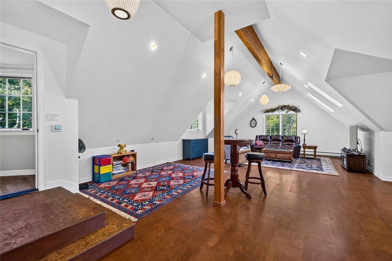This spacious, open-concept attic room features vaulted ceilings with exposed wooden beams and multiple pendant light fixtures. The area is divided into a play space with a colorful rug and a cozy seating area with a leather sofa, all set against a backdrop of white walls and warm-toned flooring. Large windows and skylights allow for ample natural light, creating an airy and versatile living space.