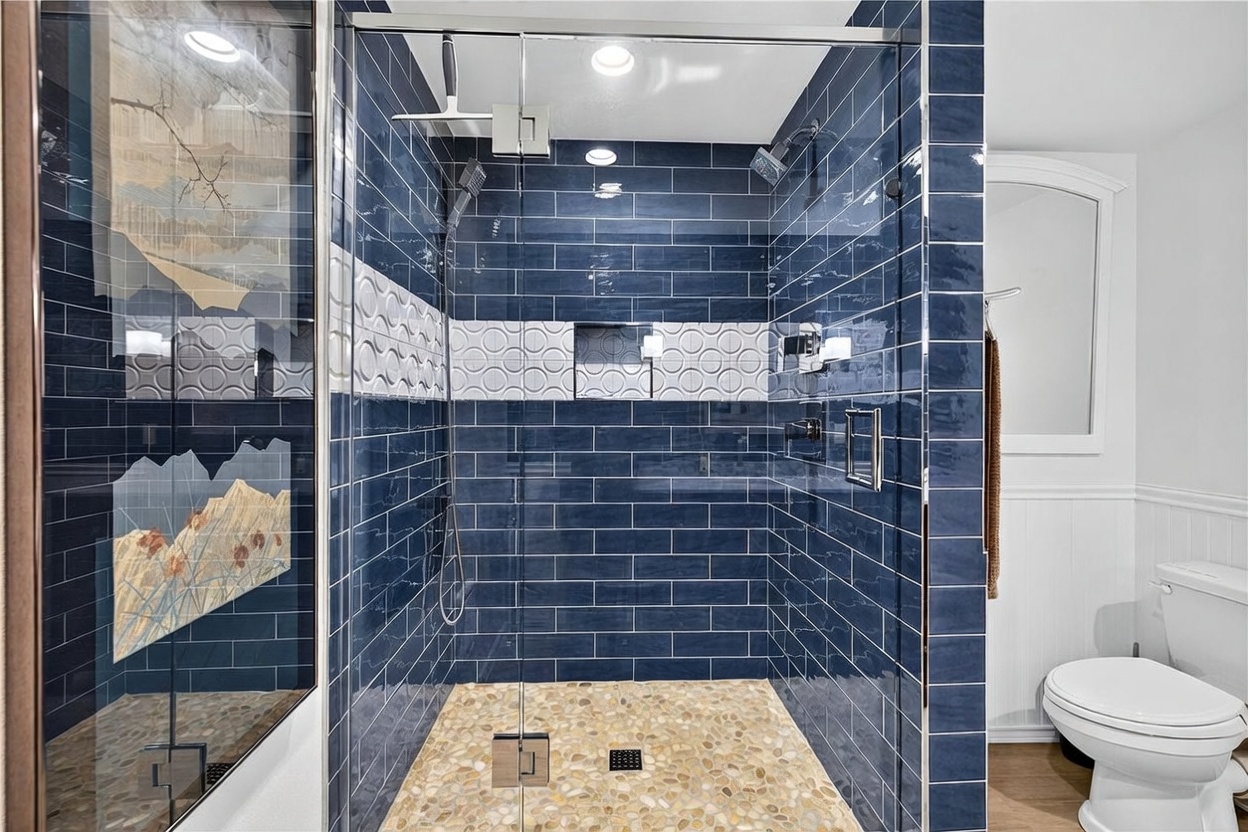 This bathroom features a walk-in shower enclosed by glass panels, showcasing striking deep blue subway tiles accented by a decorative band of circular patterned tiles. The shower floor is finished with natural river stones, and the space includes both a rainfall showerhead and a handheld sprayer. To the right, a white toilet is positioned against a wall with beadboard wainscoting, creating a clean and contrasting aesthetic.