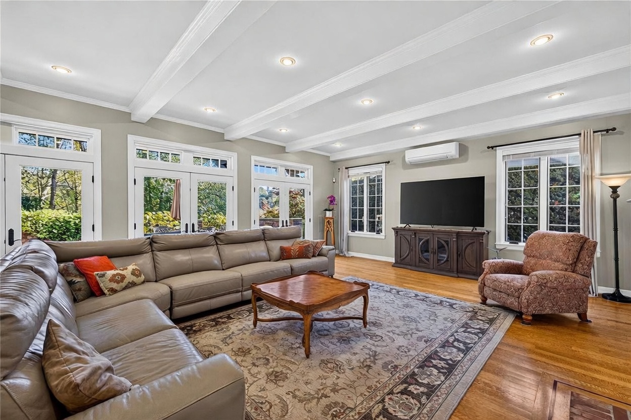 This spacious living room features a large, neutral-toned sectional sofa arranged around a central wooden coffee table on a patterned area rug. The room is brightened by multiple sets of French doors and windows that provide ample natural light and views of the outdoors. A wall-mounted television sits above a dark wood media console, while the ceiling is accented with decorative beams and recessed lighting.