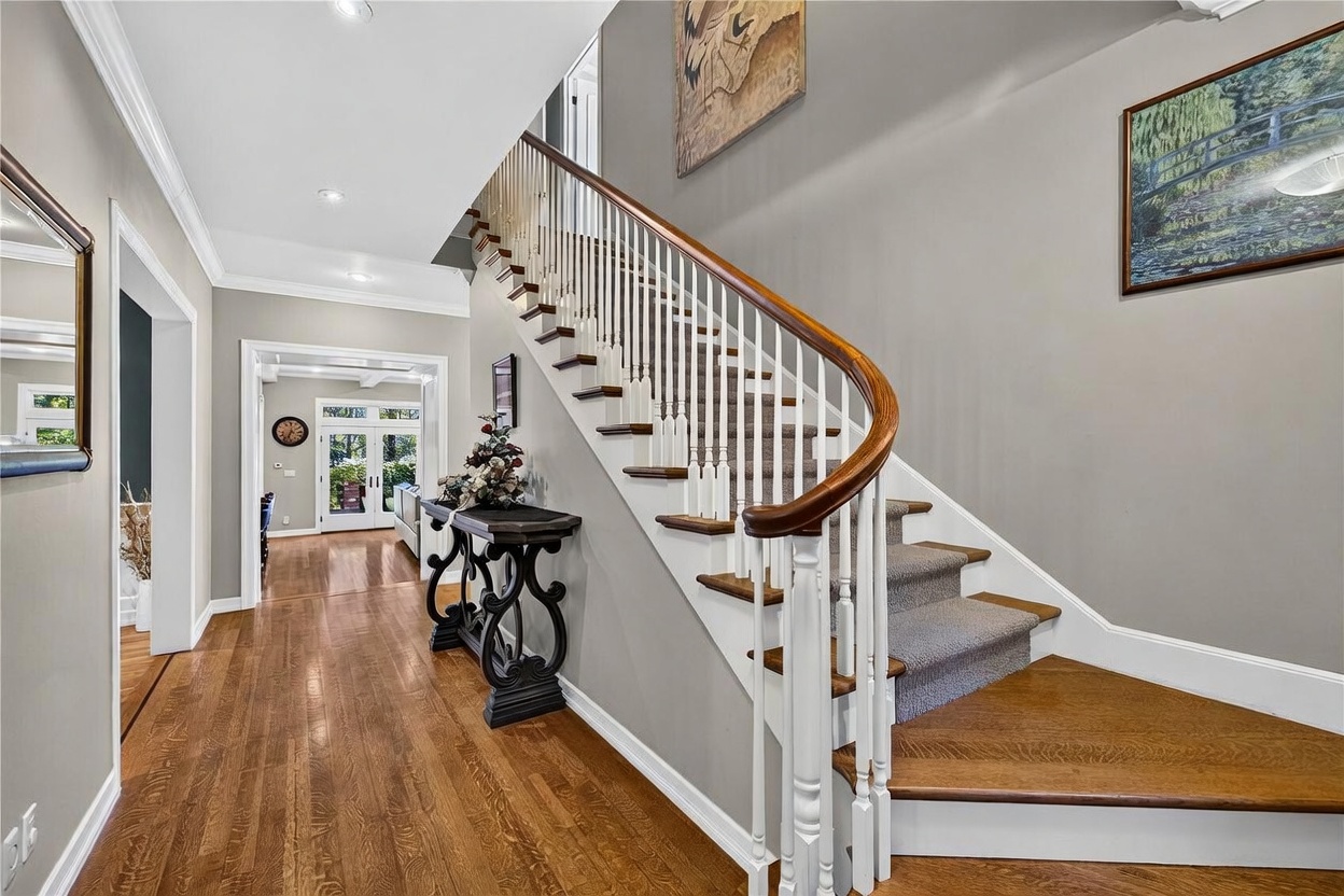 This elegant foyer features a grand staircase with a curved wooden handrail and white balusters, set against soft gray walls. The space is anchored by warm-toned hardwood flooring and a decorative console table adorned with a floral arrangement. A clear line of sight leads through an arched doorway into an adjacent room, creating an inviting and open atmosphere.