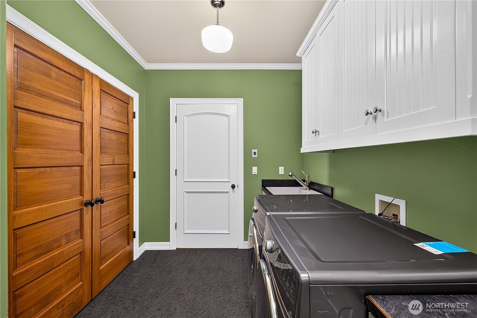 This functional laundry room features dark gray appliances positioned beneath white upper cabinetry and a small utility sink. The space is painted in a muted green tone, complemented by warm-toned wooden double doors on the left and a white interior door in the center. A single pendant light fixture hangs from the ceiling, illuminating the dark flooring and providing a clean, organized utility area.