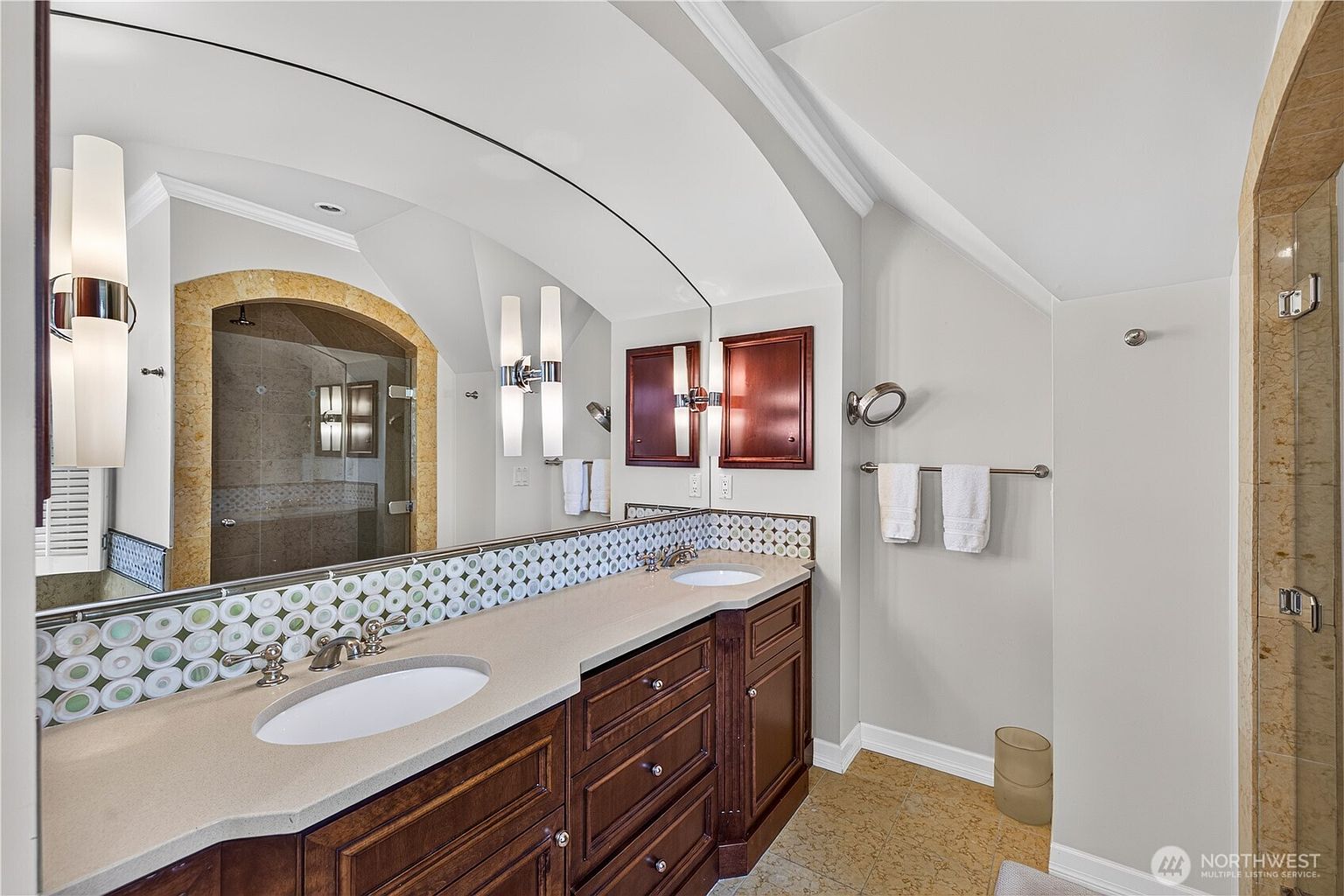 This primary bathroom features a dual-sink vanity with dark wood cabinetry and a light-colored stone countertop. A large, arched mirror spans the length of the vanity, reflecting the shower area which is enclosed by a glass door and framed with decorative stone tile. The space is accented by a band of circular mosaic tile above the vanity and includes wall-mounted light fixtures.