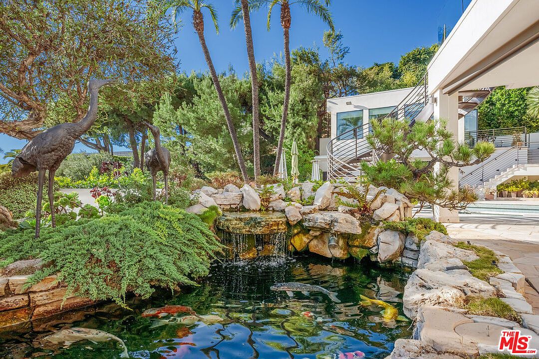 This serene outdoor space features a tiered rock waterfall cascading into a koi pond filled with colorful fish. The garden is accented by two bronze crane statues, lush greenery, and tall palm trees, creating a tranquil, resort-like atmosphere. A modern home with a spiral staircase and large windows is visible in the background, seamlessly blending the architecture with the natural landscape.
