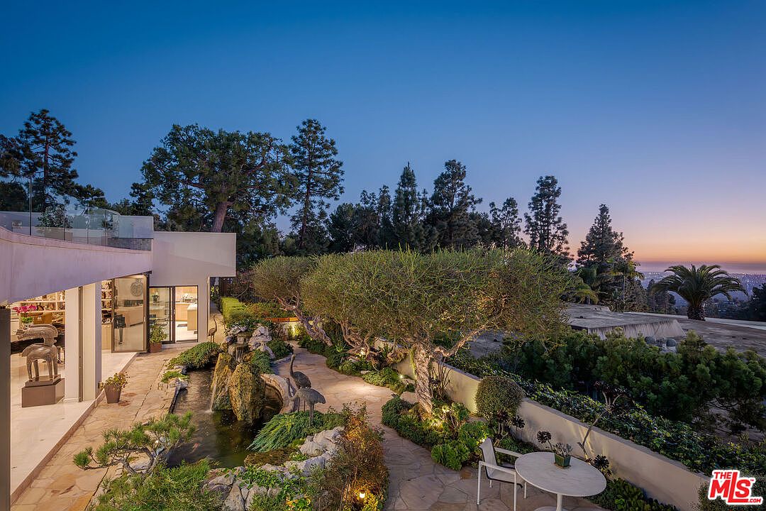 This serene outdoor space features a meticulously landscaped garden with a stone-lined koi pond and a cascading water feature. A winding stone pathway leads through the lush greenery, complemented by mature trees and soft landscape lighting that creates an inviting evening ambiance. The scene is framed by a modern home with large glass doors, offering a seamless transition between indoor and outdoor living with a panoramic view of the city lights in the distance.