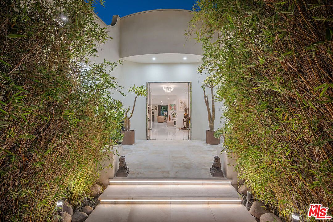 This striking entryway features a grand, illuminated staircase leading to a modern, open-concept home interior. The path is framed by lush, tall bamboo stalks on either side, creating a private and serene atmosphere, while two stone lion statues flank the entrance. The perspective is centered and symmetrical, drawing the eye through the open glass doors into the bright, gallery-like living space beyond.