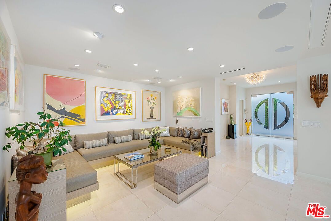 This spacious, contemporary living room features a large, L-shaped sectional sofa in a neutral tone, accented by a glass-topped coffee table and a matching ottoman. The walls are adorned with vibrant, colorful abstract artwork, and the room is illuminated by numerous recessed ceiling lights. A wide entryway with glass double doors featuring a prominent circular logo leads into the space, creating an open and airy atmosphere with polished light-colored flooring throughout.
