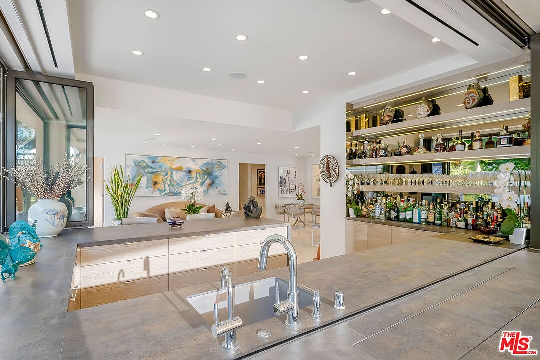 This modern kitchen features a large central island with a matte-finish countertop and a sleek, integrated sink. Behind the island, a mirrored bar area with illuminated shelving displays glassware and decorative masks, creating a sophisticated entertaining space. The open-concept design flows into a bright living area adorned with contemporary artwork and light-toned furniture, offering a seamless transition between rooms.
