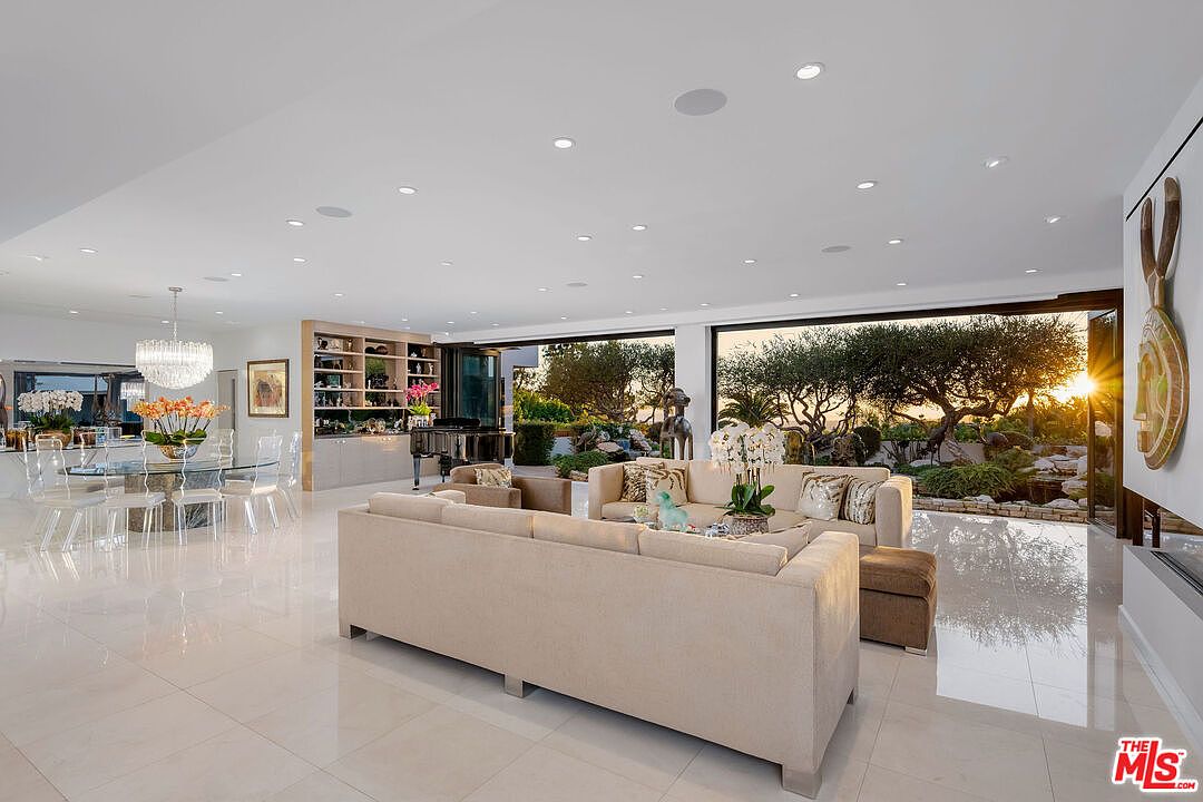 This expansive, open-concept living and dining area features polished light-colored flooring and a neutral color palette that enhances the sense of space. A large, plush sofa serves as the centerpiece of the living area, which transitions seamlessly into a dining space with a glass-topped table and modern chairs. The room is defined by massive glass walls that slide open to reveal a sunset view of the outdoor landscape, creating a fluid connection between the interior and the exterior.