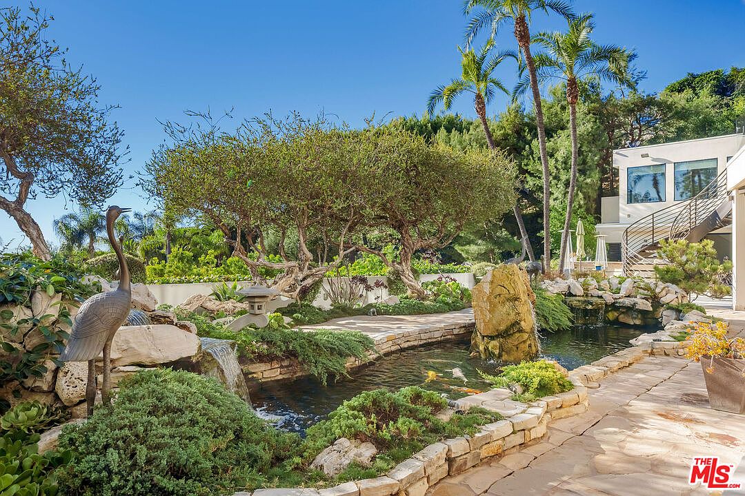 This serene backyard features a meticulously landscaped koi pond with a stone waterfall and a decorative heron statue. The garden is accented by mature trees, lush greenery, and stone pathways that lead toward a modern home with a curved staircase. The overall atmosphere is tranquil and reminiscent of a private Japanese-inspired retreat.