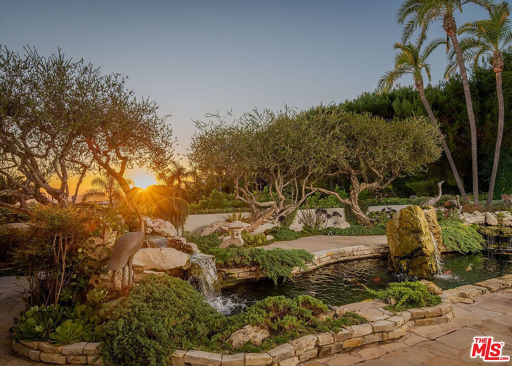 This serene backyard garden features a tiered stone koi pond with a cascading waterfall and a prominent rock feature. The landscape is accented by mature, twisted trees, lush greenery, and decorative crane statues, all set against a warm, golden sunset. The scene is captured from a low, eye-level perspective, emphasizing the tranquil atmosphere and the intricate stonework of the pond's edge.