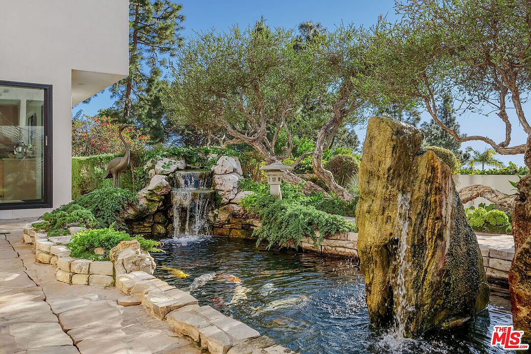 This serene backyard garden features a multi-tiered koi pond with a naturalistic waterfall cascading over stacked stone. A large, weathered boulder serves as a focal point in the foreground, while lush greenery and mature trees provide a tranquil, shaded atmosphere. The scene is captured from a ground-level perspective, highlighting the seamless integration of the water feature with the surrounding landscape and the adjacent home's exterior wall.
