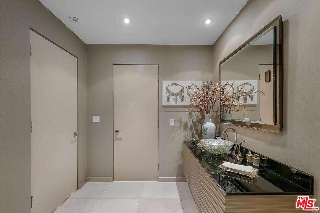 This contemporary powder room features a sleek vanity with a dark stone countertop and a unique, textured vessel sink. A large, framed mirror hangs above the vanity, reflecting the room's neutral color palette and decorative wall art. The space is illuminated by recessed ceiling lights, creating a clean and sophisticated atmosphere.