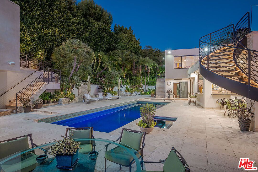 This evening shot captures a luxurious backyard oasis featuring a long, rectangular swimming pool and an integrated spa. The patio area is paved with light-colored stone and includes a dining set in the foreground, while a striking spiral staircase leads to an upper level. Lush landscaping, including mature trees and palms, frames the property, creating a private and serene atmosphere.