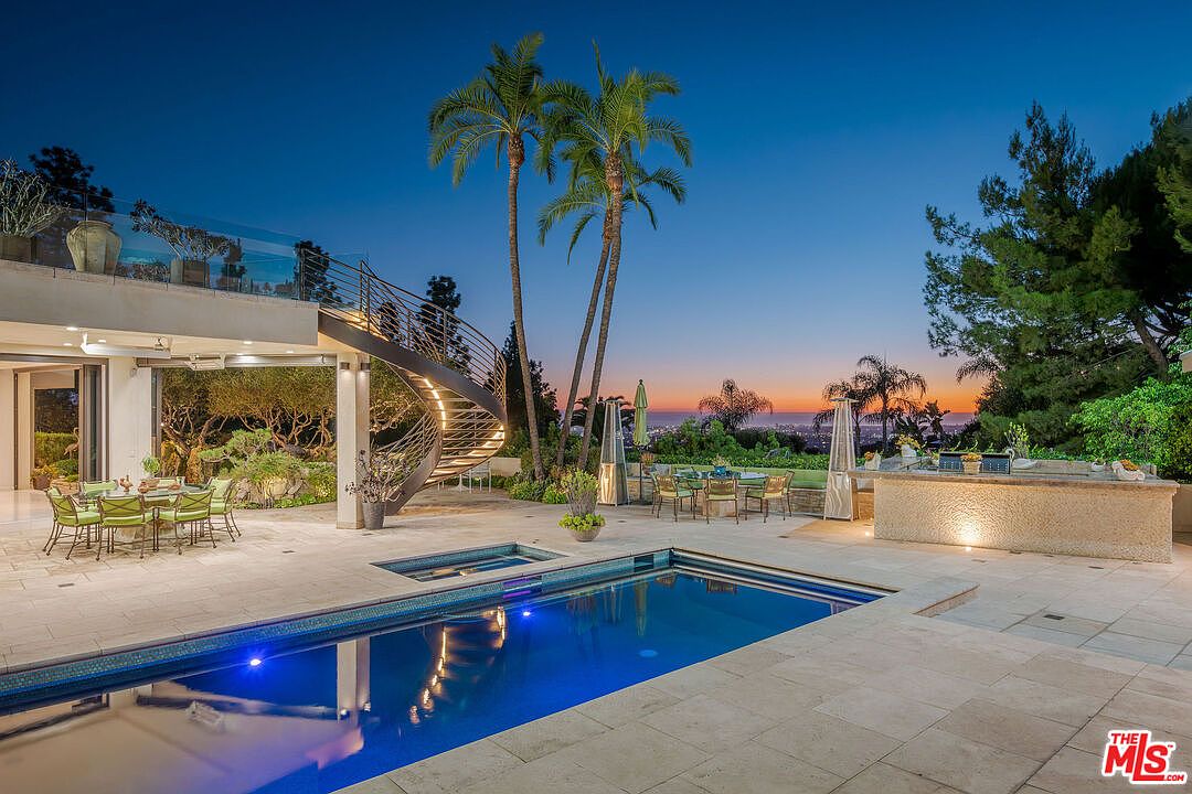 This luxurious outdoor living space features a pristine swimming pool and integrated spa set against a stunning twilight sky. A striking spiral staircase connects the patio to an upper-level balcony, while a built-in outdoor kitchen and multiple dining areas provide ample space for entertaining. The scene is framed by tall palm trees and lush landscaping, offering a sophisticated and serene atmosphere with a panoramic view of the city lights in the distance.
