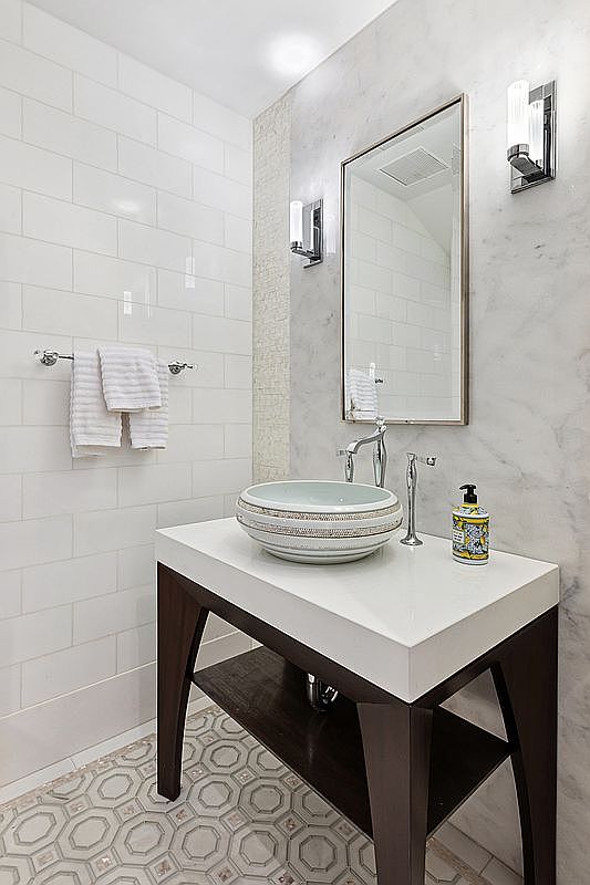 This elegant powder room features a vessel sink perched atop a dark wood vanity with a lower shelf. The walls are adorned with a combination of white subway tile and light-toned stone, complemented by a rectangular mirror and modern wall-mounted light fixtures. The floor is finished with a decorative, geometric patterned tile, creating a sophisticated and polished aesthetic.