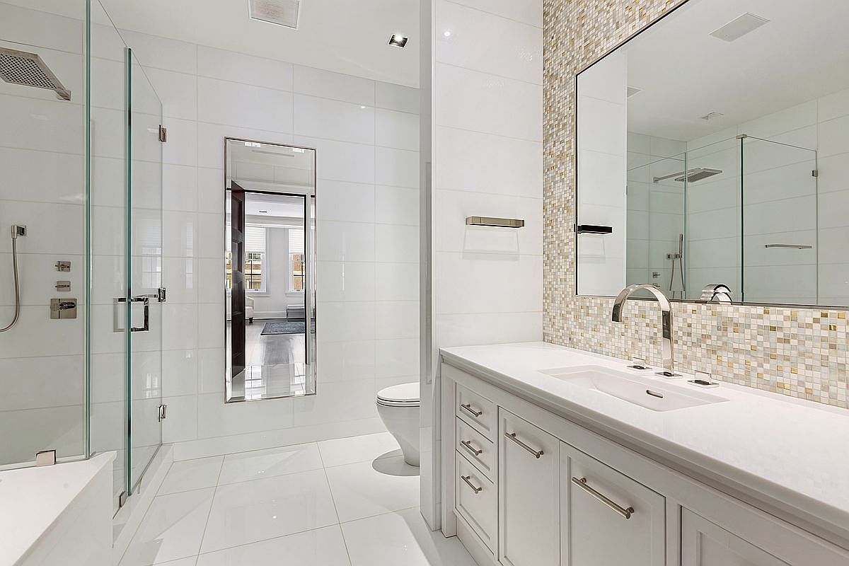 This modern primary bathroom features a sleek white vanity with a stone countertop and a large mirror reflecting a mosaic-tiled accent wall. To the left, a glass-enclosed walk-in shower with polished fixtures provides a clean, open aesthetic, while a tall, narrow mirror reflects the adjacent room. The space is finished with bright white wall and floor tiles, creating a crisp, high-end atmosphere.