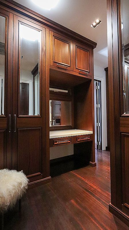 This elegant walk-in closet features rich, dark-toned wood cabinetry with integrated mirror panels and a dedicated vanity area. The space is finished with polished dark wood flooring and a plush, textured stool in the foreground, creating a sophisticated and organized atmosphere. The perspective looks directly into the vanity nook, highlighting the custom millwork and recessed lighting.