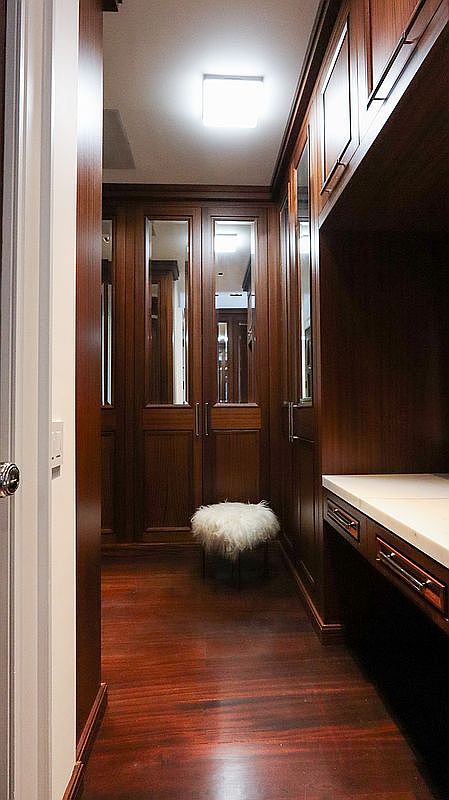 This elegant walk-in closet features rich, dark wood cabinetry with integrated mirrored doors and a built-in vanity area with a light-toned countertop. A plush, white faux-fur stool sits in the center of the room, adding a touch of luxury to the warm, polished wood flooring. The perspective is a straight-on view looking down the narrow corridor, highlighting the sophisticated storage solutions and refined craftsmanship.
