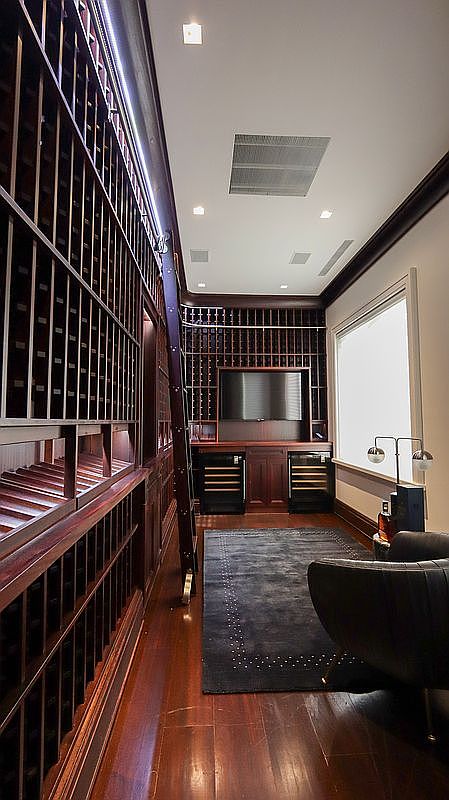This sophisticated wine cellar features extensive custom wood shelving that spans the walls, providing ample storage for a large collection. A rolling library-style ladder allows access to the upper racks, while the back wall incorporates a built-in television above two under-counter wine refrigerators. A dark area rug centers the room, complemented by a comfortable leather armchair and a modern lamp, creating a refined space for tasting and relaxation.