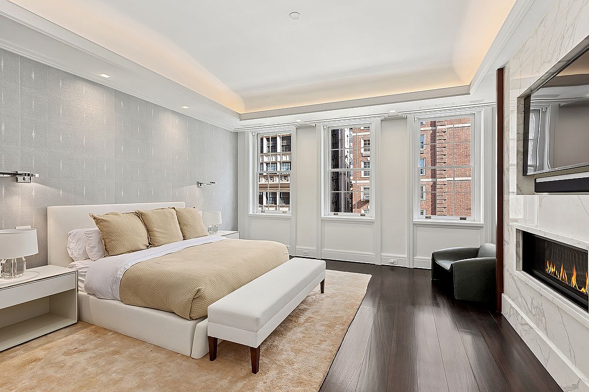 This elegant primary bedroom features a neutral color palette with a plush bed centered against a textured, light-colored accent wall. The room is illuminated by natural light streaming through three tall windows, while a modern linear fireplace is integrated into a stone-clad wall opposite the bed. Dark wood flooring and a soft area rug anchor the space, creating a sophisticated and tranquil atmosphere.