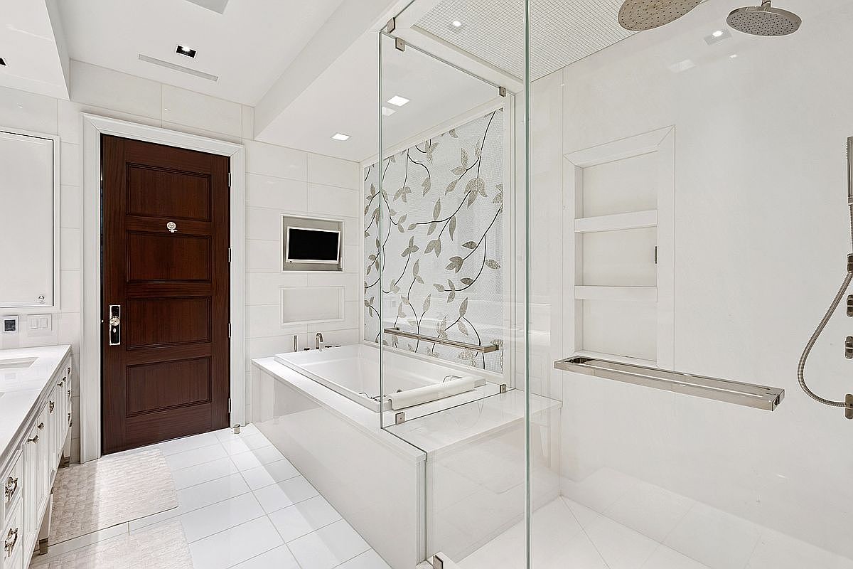 This luxurious primary bathroom features a seamless glass-enclosed walk-in shower and a deep soaking tub set against a decorative leaf-patterned tile accent wall. The space is finished with bright white wall tiles, a dark wood door, and a sleek vanity area, creating a clean and sophisticated atmosphere. The perspective captures the entire wet room layout, highlighting the modern fixtures and the integrated television above the tub.