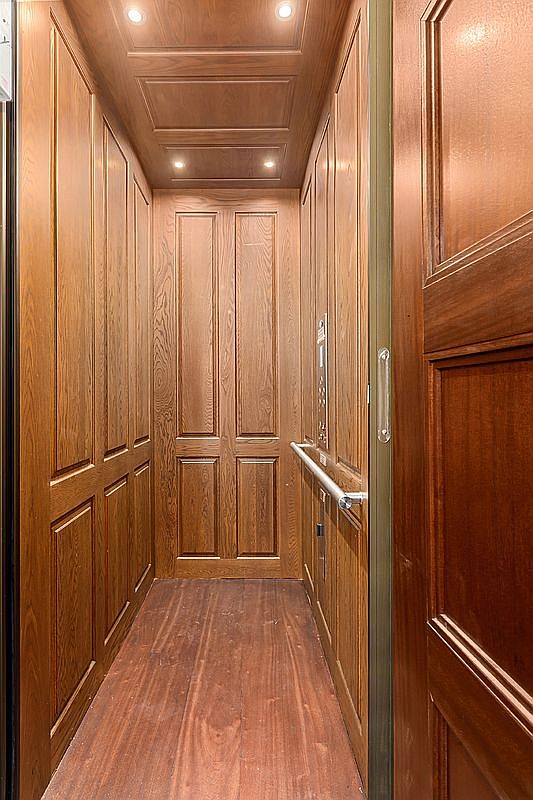 This image shows the interior of a private elevator finished with rich, warm-toned wood paneling throughout the walls and ceiling. A metal handrail runs along the side wall, and a control panel is mounted near the entrance, creating a sophisticated and classic aesthetic. The perspective is a straight-on view looking into the narrow, enclosed space, emphasizing the high-quality craftsmanship and luxurious feel of the elevator cabin.