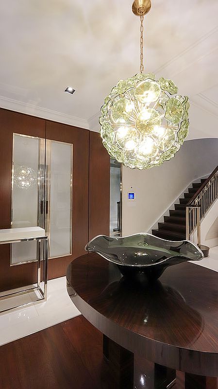 This elegant entryway features a dark wood oval table topped with a large, dark glass bowl, positioned beneath a striking, multi-faceted glass pendant chandelier. To the left, a sleek console table sits against a wall of dark wood paneling with integrated frosted glass doors, while a staircase with a metal railing ascends in the background. The perspective is eye-level, capturing a sophisticated and polished foyer that blends modern luxury with warm, rich tones.