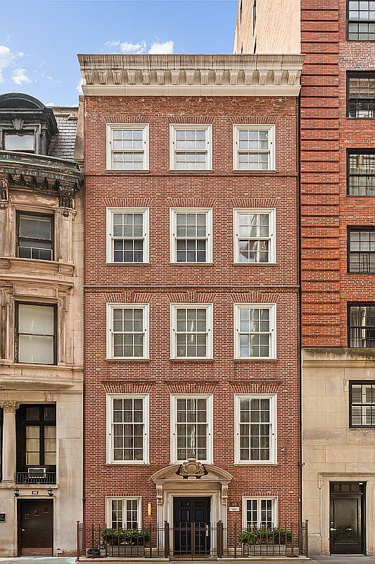 This image presents a direct, eye-level front view of a classic, multi-story brick townhouse in an urban setting. The facade features symmetrical rows of multi-pane windows, a decorative cornice at the roofline, and a formal, centered entryway with a pedimented surround. The building is flanked by neighboring structures, showcasing a traditional architectural style typical of historic city residences.