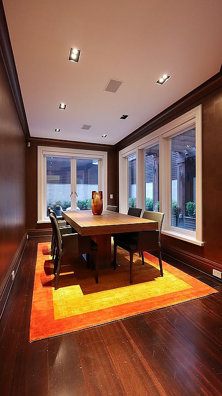 This formal dining room features dark wood paneling and rich hardwood flooring, creating a sophisticated and moody atmosphere. A large, rectangular wooden table serves as the centerpiece, surrounded by modern chairs, all set atop a vibrant orange area rug. The room is illuminated by recessed ceiling lights and benefits from natural light streaming through large windows and glass doors that look out onto an outdoor space.
