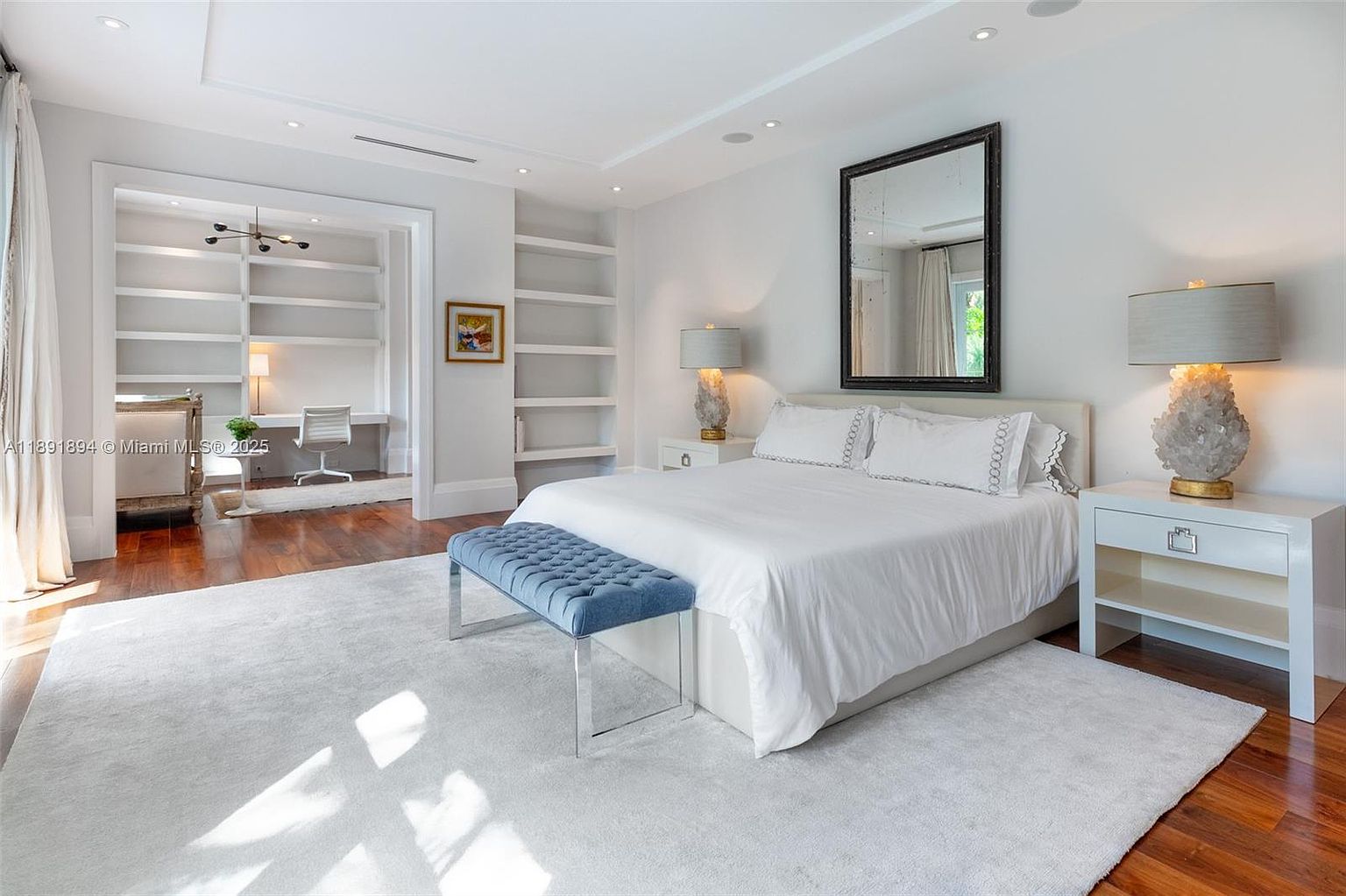 This is a bright and spacious primary bedroom featuring a large bed with white linens, a tufted blue bench, and built-in shelving. The room has hardwood floors partially covered by a light-colored rug, and a separate office nook is visible through an archway. The overall aesthetic is clean, modern, and luxurious.