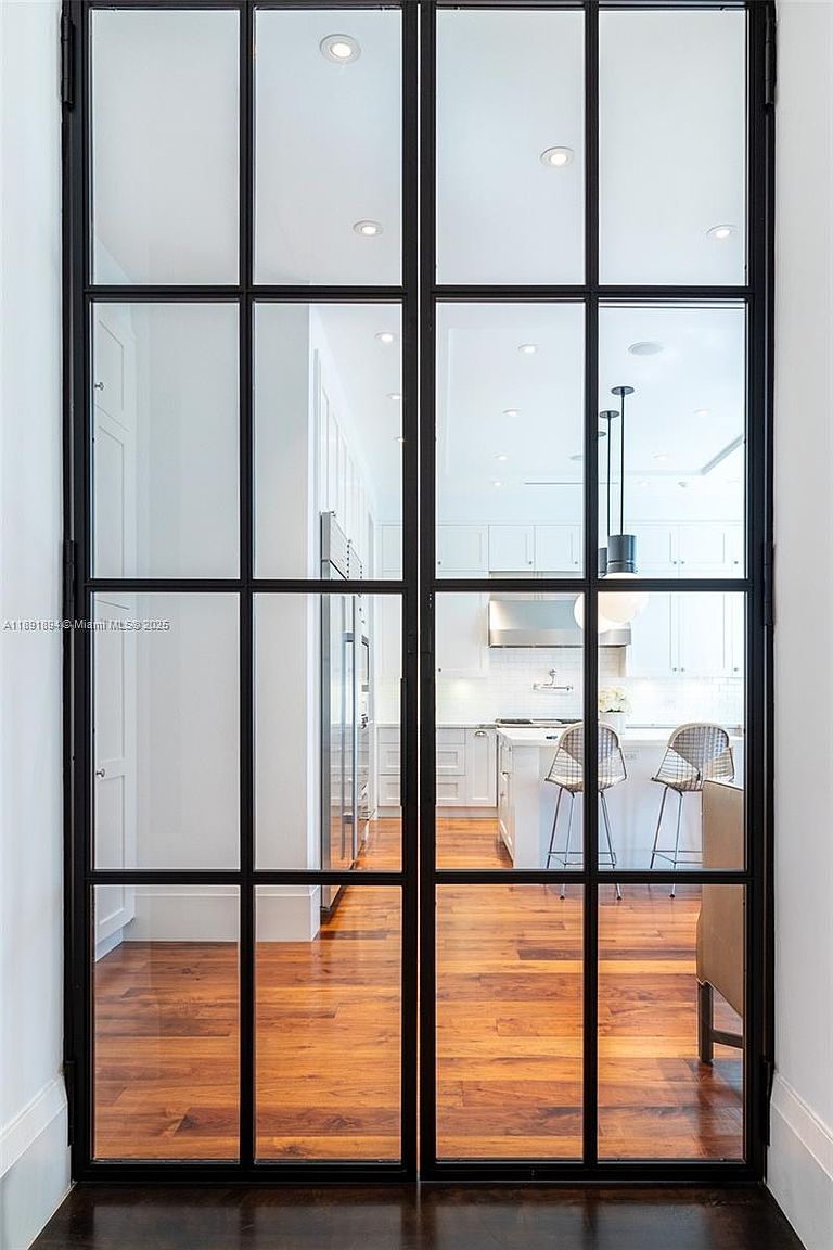 The image showcases a modern kitchen viewed through a set of black-framed glass doors. The kitchen features white cabinetry, a central island with bar stools, and stainless steel appliances. The hardwood flooring adds warmth to the space, creating an inviting and stylish atmosphere.