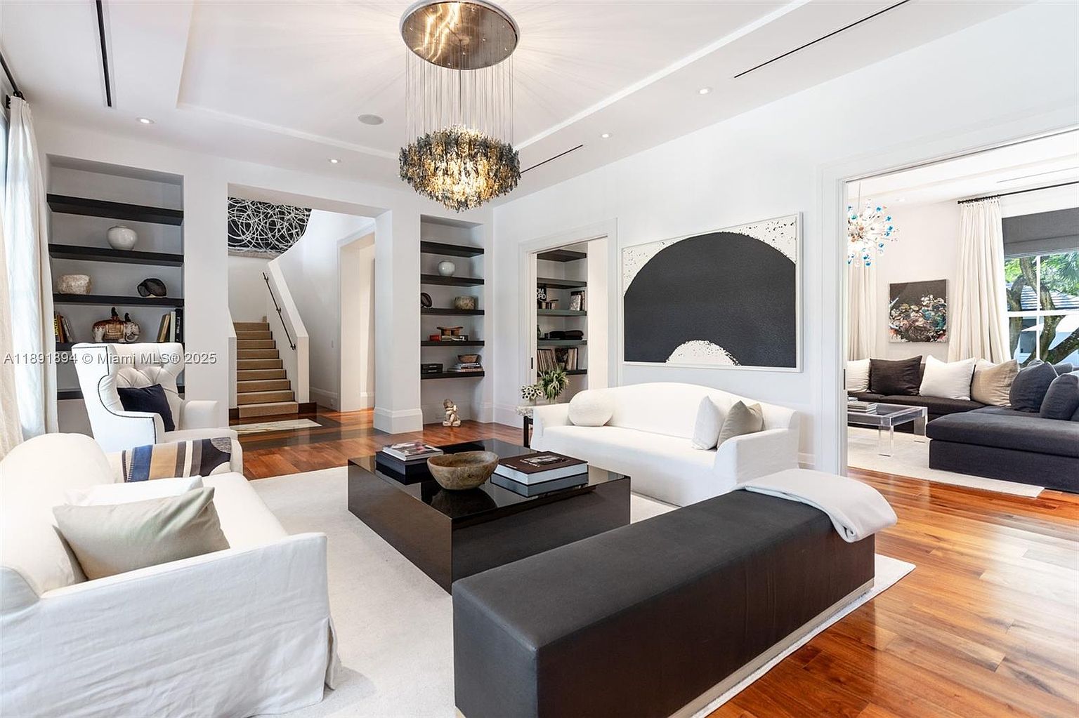 This is an interior shot of a luxurious living room featuring hardwood floors, white walls, and a high ceiling with a modern chandelier. The room is furnished with white sofas, a dark bench, and a dark coffee table, creating a sophisticated and inviting atmosphere. The perspective is from a medium angle, showcasing the depth and layout of the room, including a glimpse into an adjacent sitting area and a staircase.