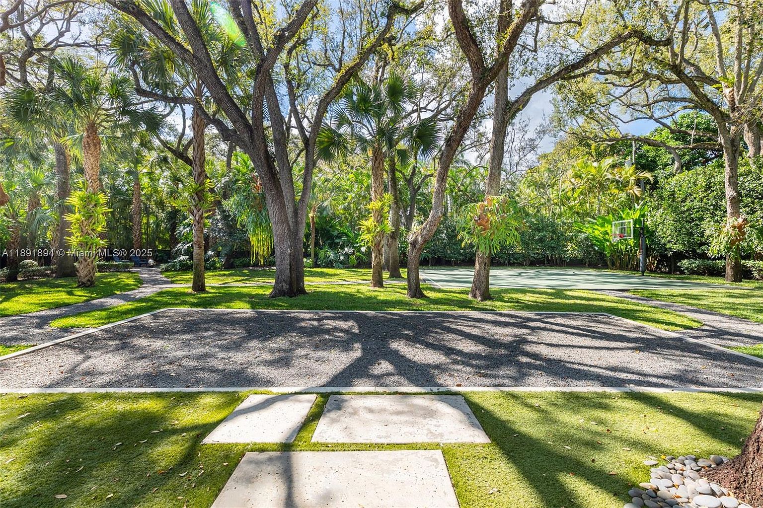 This image showcases a beautifully landscaped yard featuring mature trees, a gravel pathway, and a basketball court in the background. The foreground includes modern concrete stepping stones set in artificial turf, leading to a gravel area. The scene evokes a sense of tranquility and outdoor recreation.