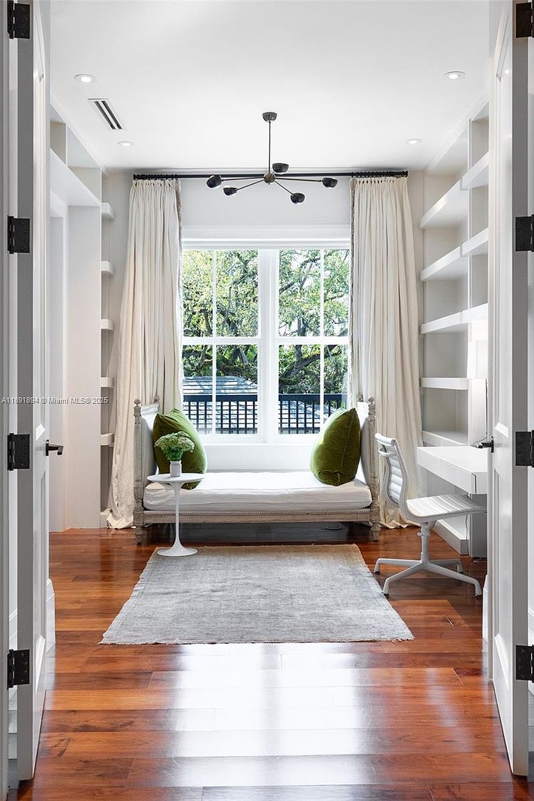 This is an interior shot of a bright and stylish office or study. The room features built-in bookshelves, a daybed with green pillows under a window, and a desk with a chair. The hardwood floors and a light-colored rug add warmth and texture to the space, creating a comfortable and functional workspace.