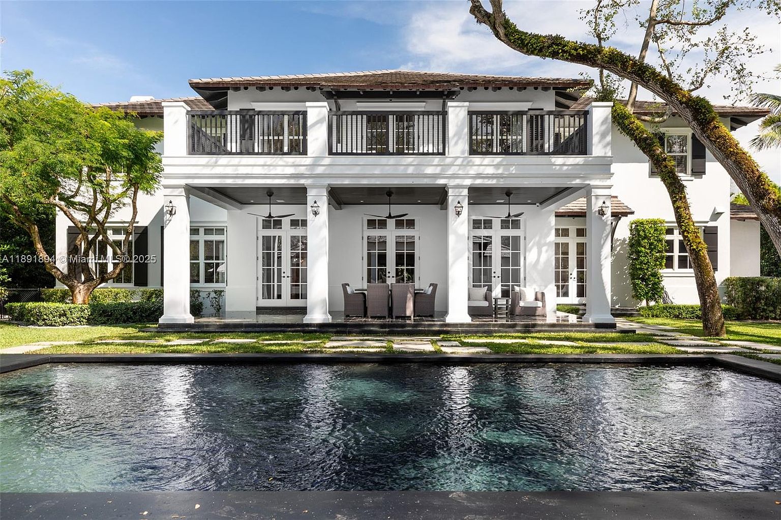 This image showcases the rear exterior of a luxurious home, emphasizing the inviting pool area. The house features a two-story design with white columns, a balcony with black railings, and multiple French doors leading to the outdoor space. The dark-colored pool reflects the house and sky, creating a serene and upscale ambiance.