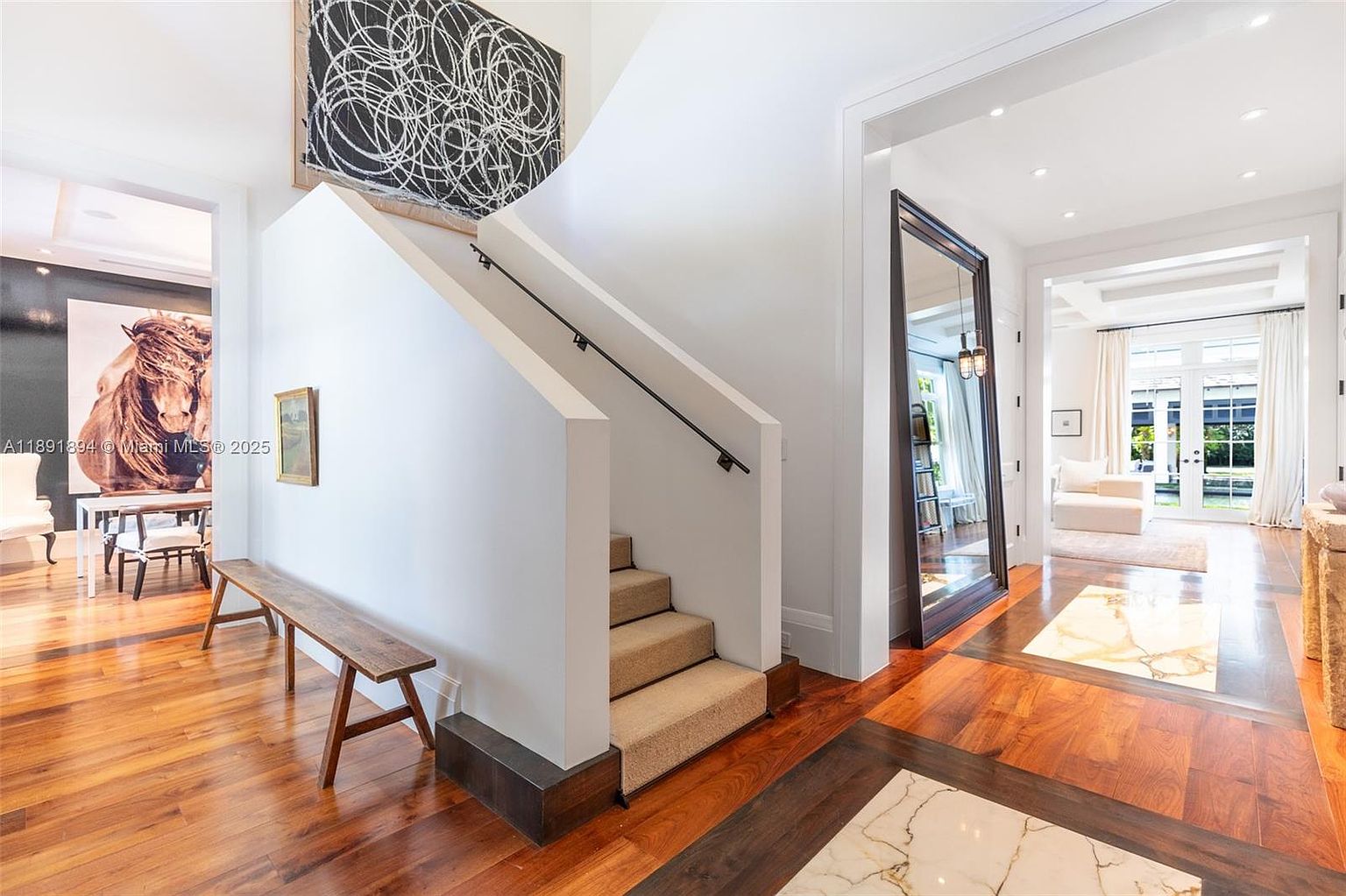 This interior shot showcases a bright and elegant hallway with a staircase. The hallway features hardwood flooring with decorative inlays, a large mirror, and access to a sunlit room. The staircase has carpeted steps and a sleek black handrail, complemented by modern artwork and a wooden bench, creating a sophisticated and inviting atmosphere.