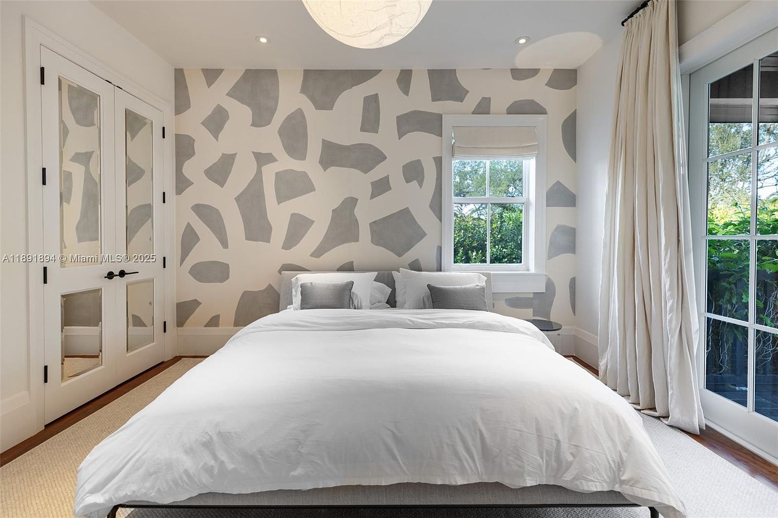 This is a bright and airy bedroom featuring a large bed with white linens, a patterned accent wall, and a window providing natural light. The room also includes a closet with mirrored doors and a door leading to the outside. The overall aesthetic is modern and inviting, creating a comfortable and stylish space.