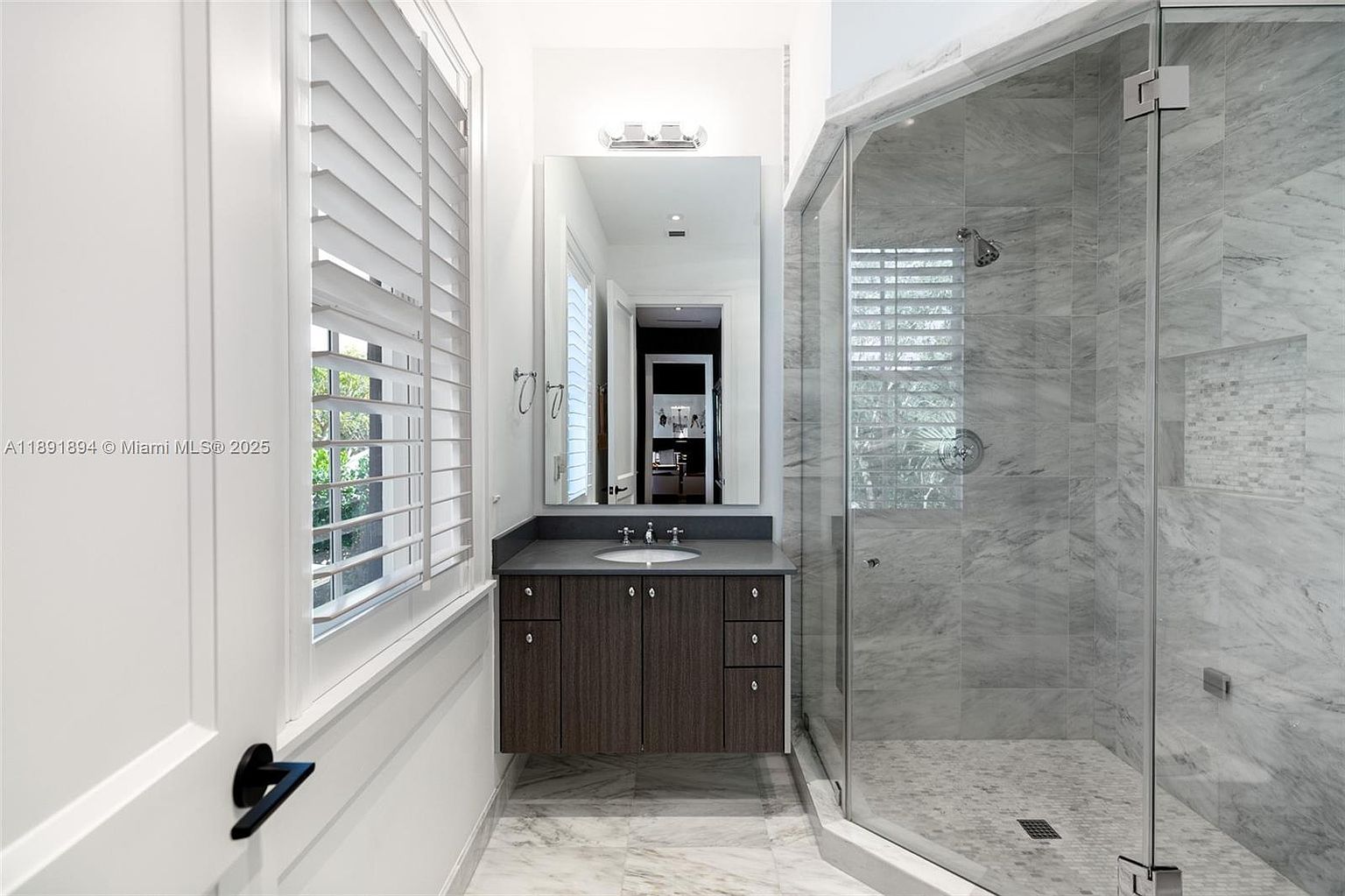 This is a well-lit bathroom featuring a modern design. The vanity has a dark wood finish with a gray countertop and a rectangular mirror above. A glass-enclosed shower with marble tiling adds a luxurious touch, and a window with white shutters provides natural light.