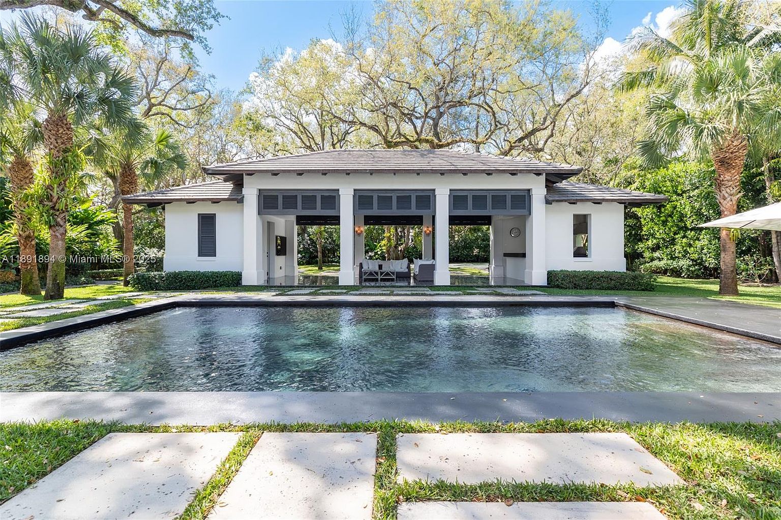 This image showcases a luxurious pool area with a modern pool house in the background. The pool features dark water and a sleek design, complemented by concrete pavers and lush green grass. The pool house has an open design with dark gray accents and white pillars, creating a sophisticated and inviting outdoor space.