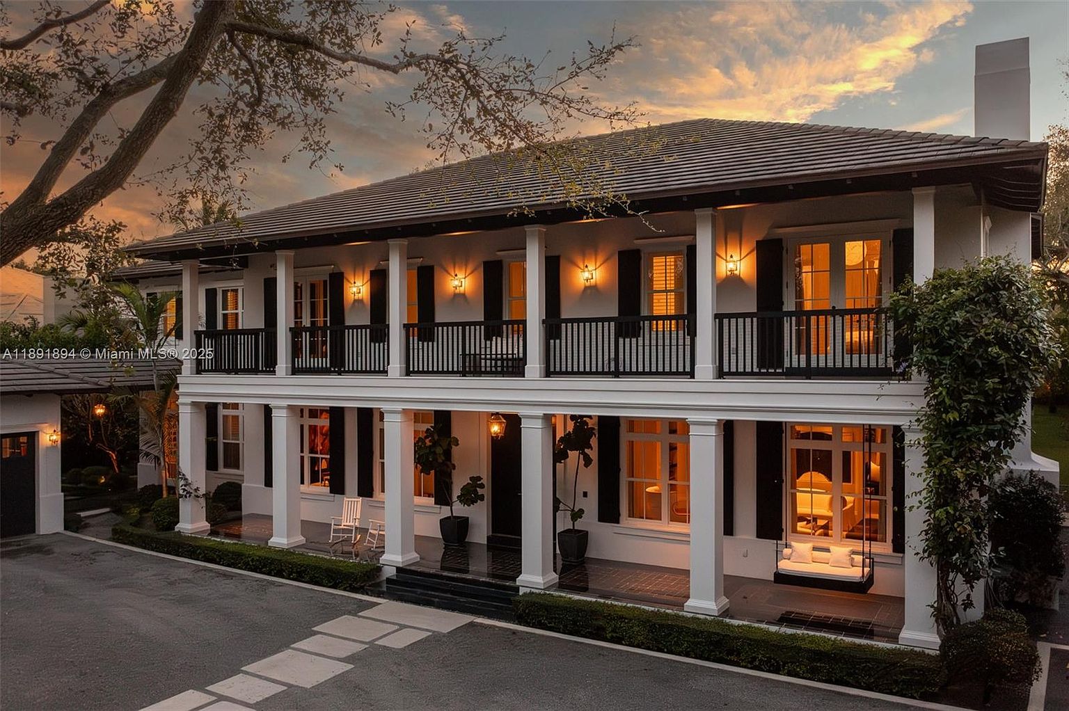 This is a front exterior view of a two-story house with a colonial style. The house features white columns supporting a balcony, black shutters, and a dark roof. The front yard is well-manicured with a paved driveway and green hedges, creating a welcoming and elegant curb appeal.