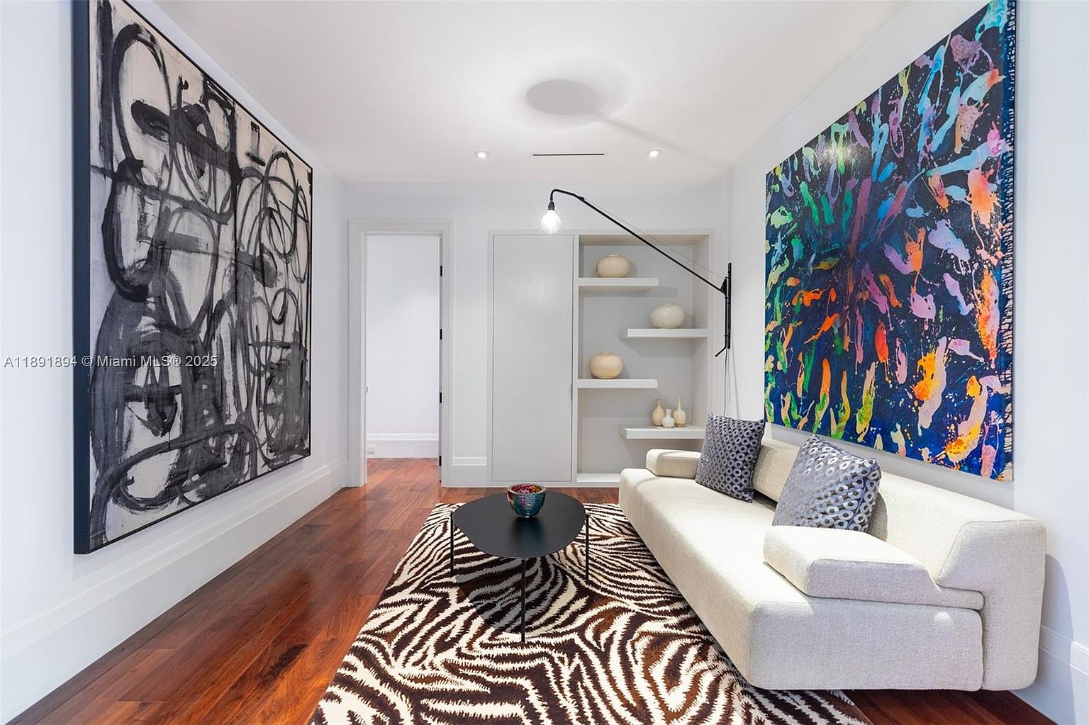 This interior shot showcases a modern living room with hardwood floors and a zebra-print rug. The room features a cream-colored sofa, two large abstract art pieces on the walls, and a black round coffee table. A built-in shelving unit with decorative vases and a sleek black wall lamp add to the room's contemporary aesthetic.