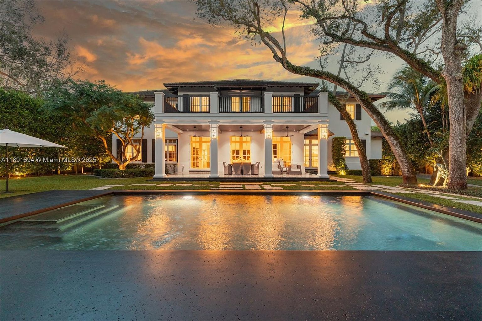 This image showcases the rear exterior of a luxurious two-story home, highlighting a stunning pool area at dusk. The pool reflects the warm glow of the sky and the house lights, creating a serene and inviting atmosphere. The home features a covered patio with columns, a second-story balcony, and lush landscaping, enhancing its curb appeal and suggesting a high-end lifestyle.