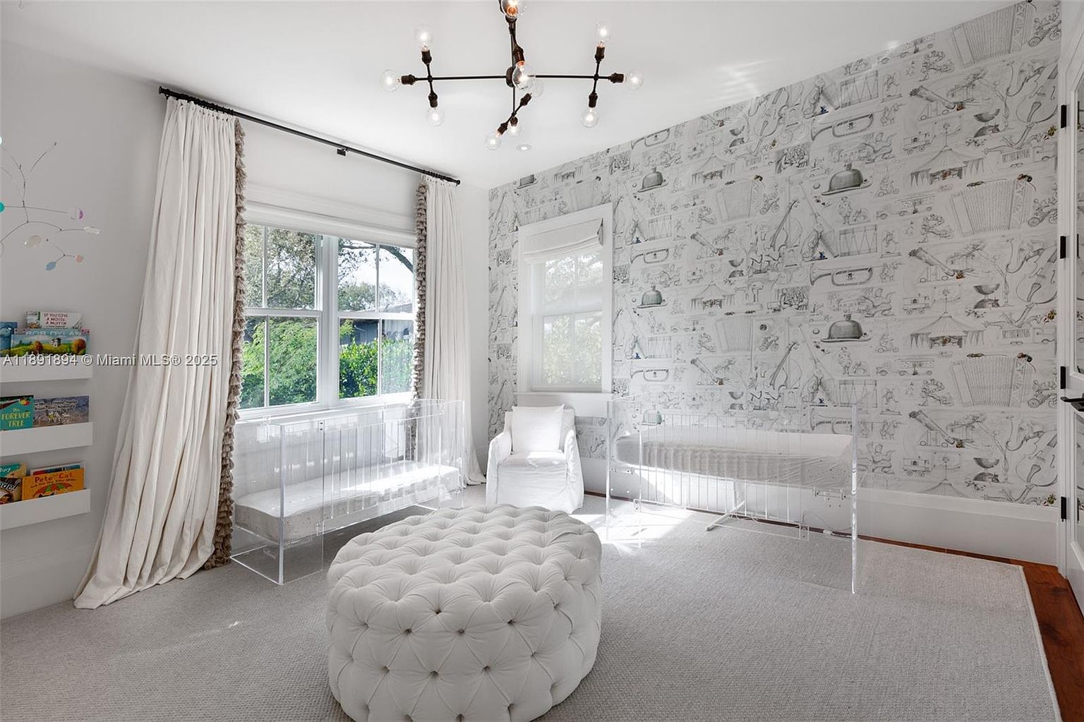 This is a bright and airy nursery featuring two clear acrylic cribs, a tufted white ottoman, and whimsical wallpaper with sketched illustrations. Natural light floods the room through two windows, complemented by elegant white curtains. The room exudes a modern and playful aesthetic, perfect for a young child.