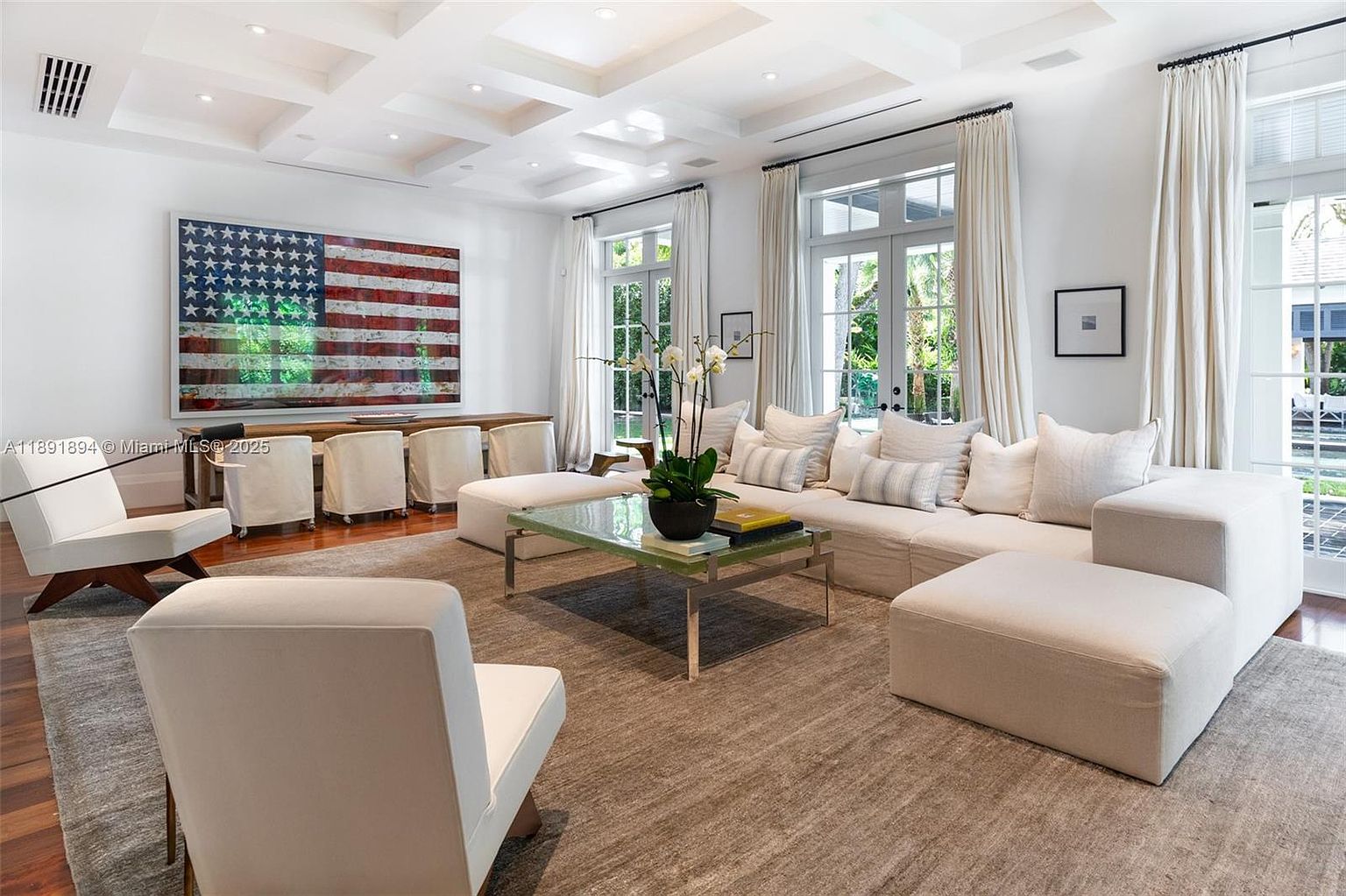 This is a bright and spacious living room featuring a large sectional sofa, a glass-topped coffee table, and two armchairs. The room is decorated in neutral tones with an American flag artwork adding a pop of color. Large windows with white curtains provide ample natural light, enhancing the airy and inviting atmosphere.